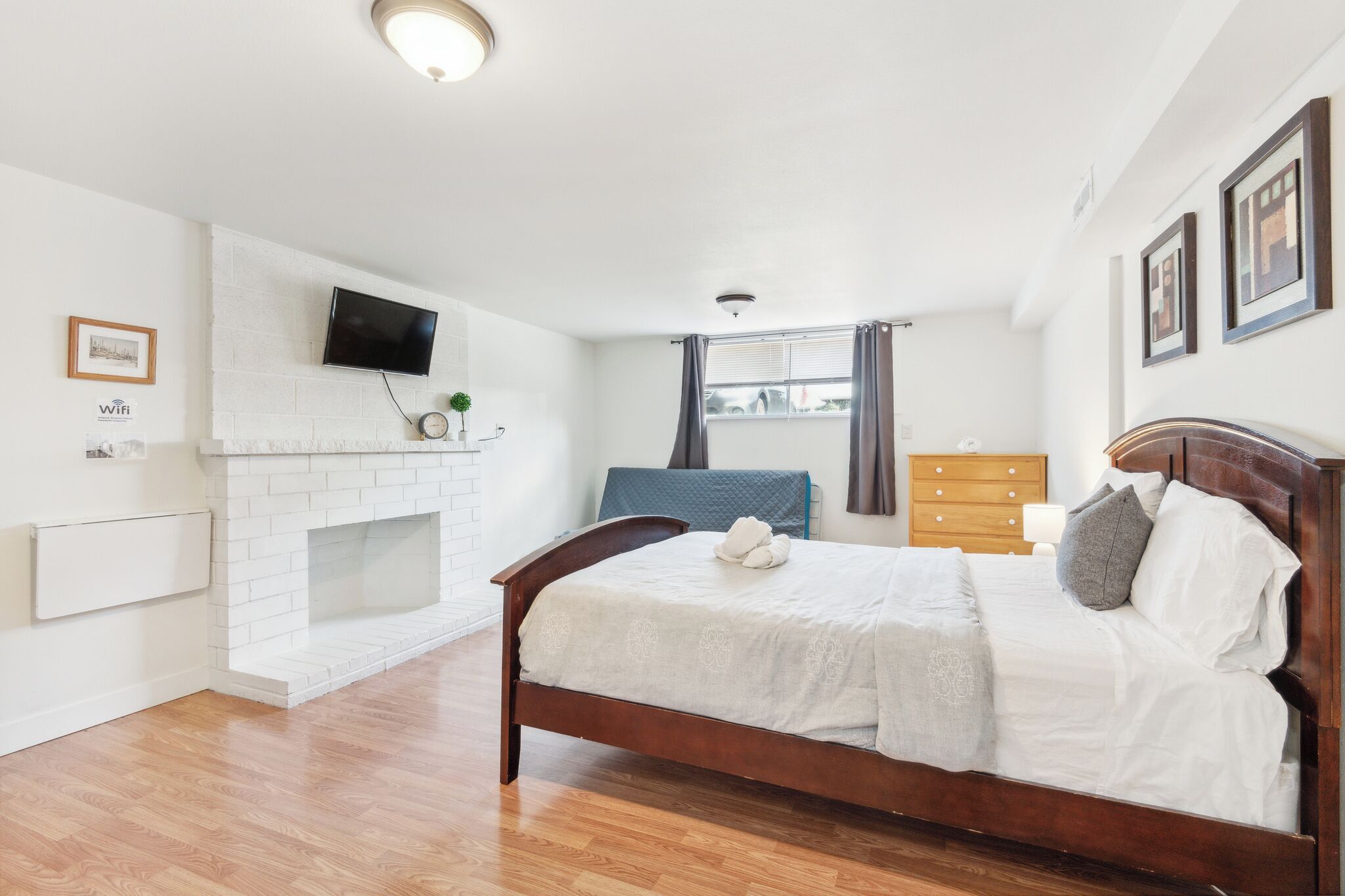 A bedroom with a wooden bed, white walls, a fireplace, a TV mounted on the wall, and a window with curtains.