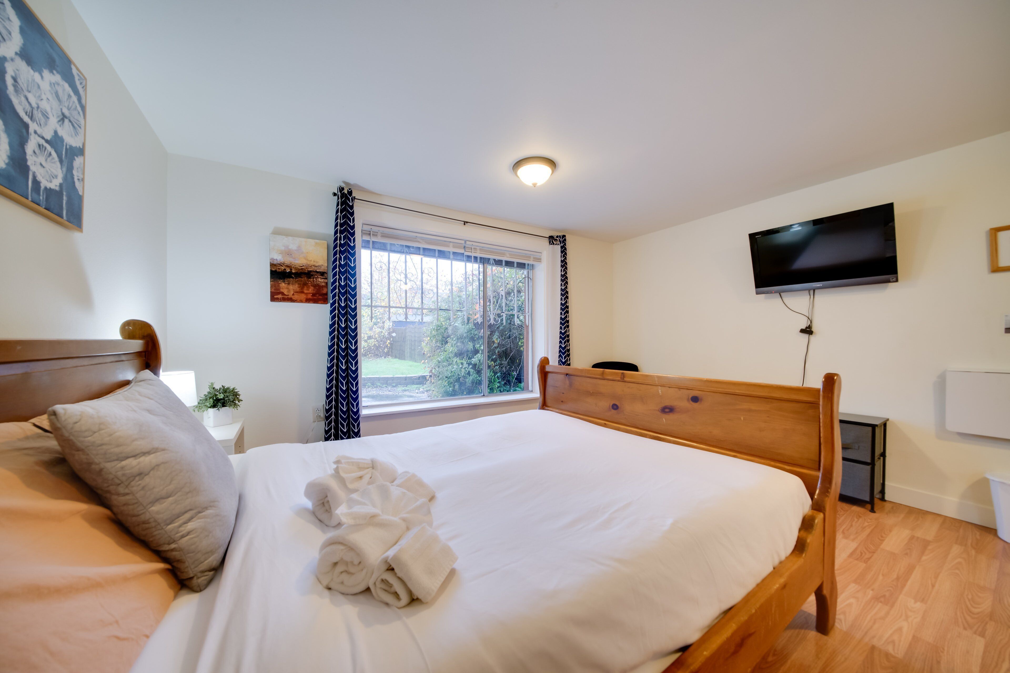 Een net opgemaakt bed met een houten hoofdbord, een flatscreen televisie aan de muur en een raam met gordijnen.