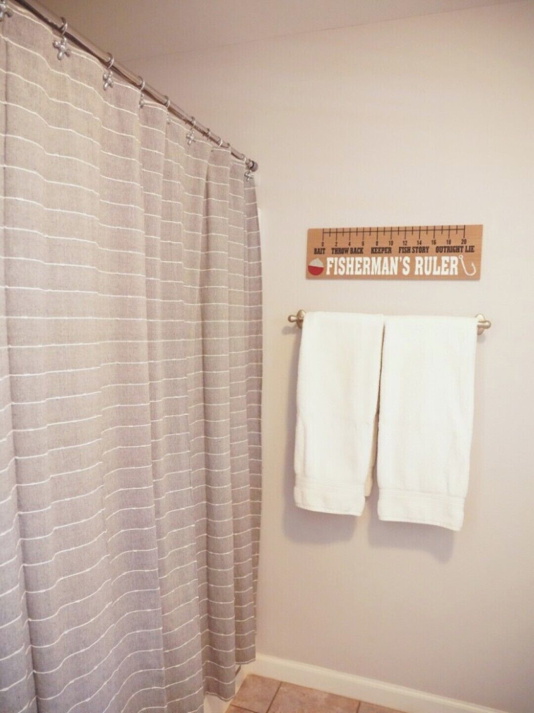 Un baño con cortina de ducha a rayas, un perchero con dos toallas blancas y un calibrador de pescador fijado a la pared.