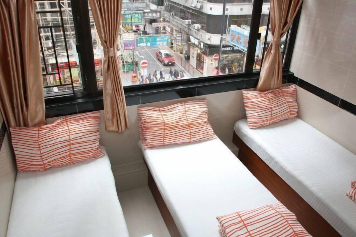 A room with three beds, each with striped pillows, a window with curtains, and a view of a busy street.