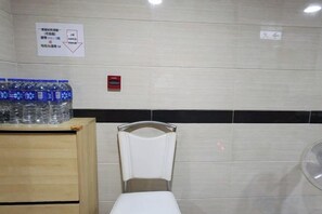 A restroom with a white toilet, a wooden cabinet with bottled water, and a wall-mounted fan. - Wing Tai Guest House (Hong Kong)