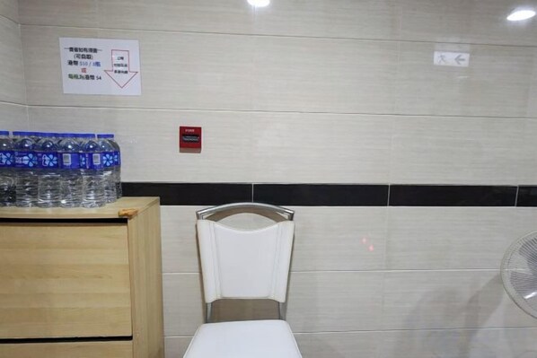 A restroom with a white toilet, a wooden cabinet with bottled water, and a wall-mounted fan. - Wing Tai Guest House (Hong Kong)