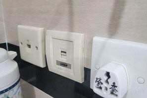 A bathroom with a black countertop, a white wall socket with a USB port, and a white electrical outlet with a plug. - Wing Tai Guest House (Hong Kong)