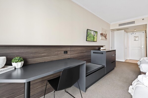 A modern hotel room with a dark wood headboard, a grey desk, a black chair, a small potted plant, and a white bed.