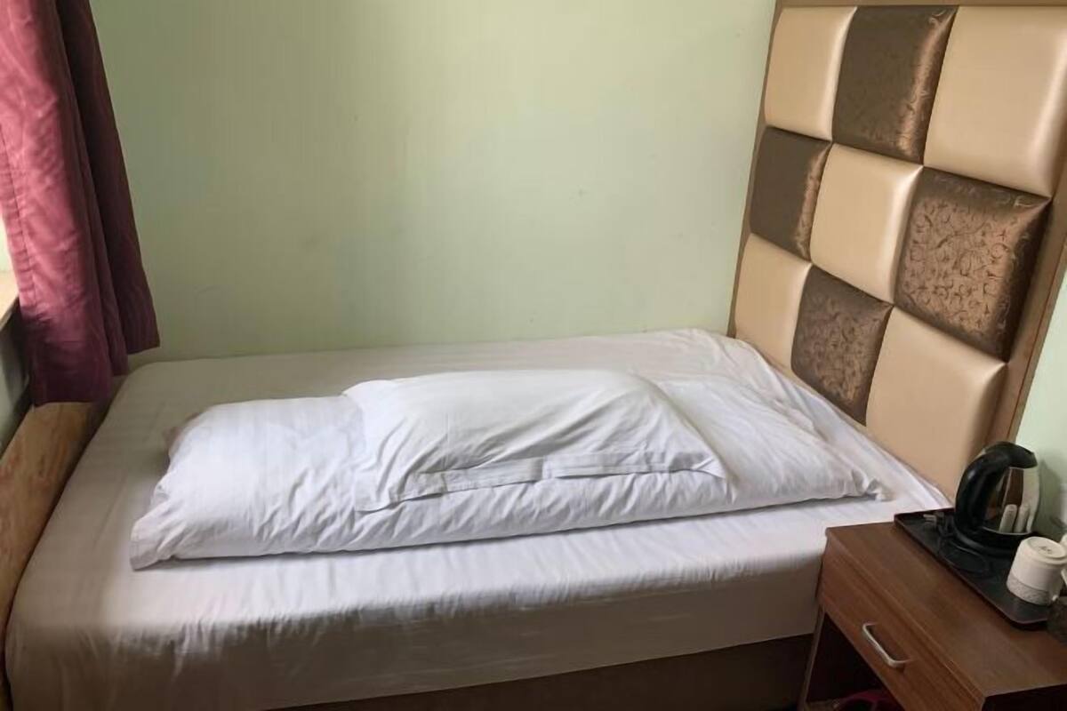A single bed with a headboard, a wooden nightstand with a kettle and a cup, and a plain wall.