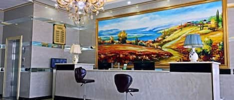 A reception area with a chandelier, a desk, two chairs, and a large landscape painting on the wall.