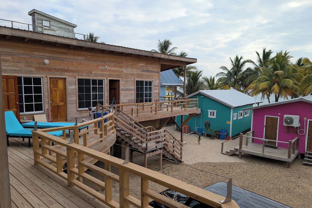 A wooden deck with a wooden staircase leading to a building with multiple rooms, including a room with a blue bed and another with a pink wall.
