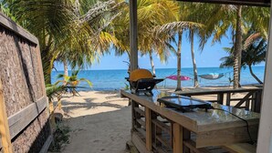 A wooden deck with a grill and a table, overlooking a sandy beach with palm trees and the ocean. - CobiaBeach.com (Hopkins)