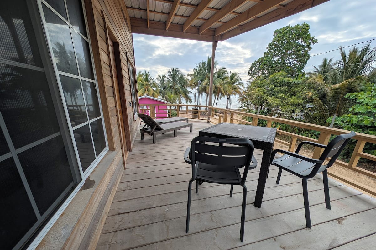 A wooden deck with a table and chairs, overlooking a beach with palm trees and a pink building in the background.