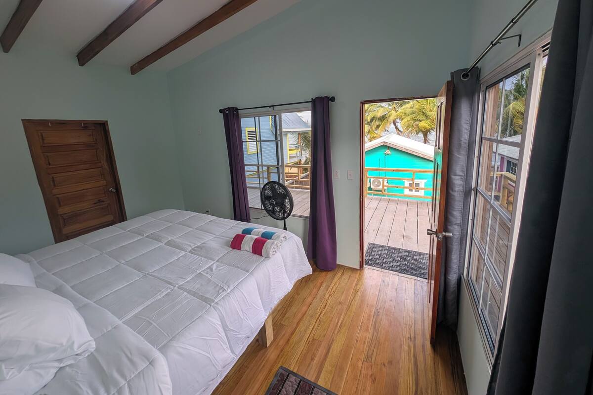 A bedroom with a bed, wooden floor, ceiling fan, and a view of a blue house and palm trees through a door.