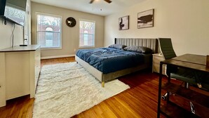 A bedroom with a bed, a desk, a chair, a ceiling fan, and two windows with blinds.