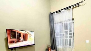Ein Hotelzimmer mit an der Wand montiertem Fernseher, einem kleinen runden Tisch mit Flasche und Speisekarte, einer Minikühlschrank und einem Fenster mit Vorhang.