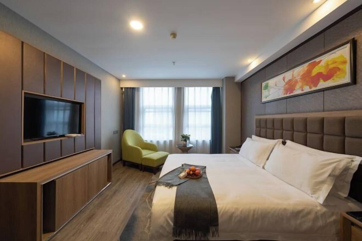 Una habitación de hotel con cama, televisión, una silla verde y ventana con cortinas.