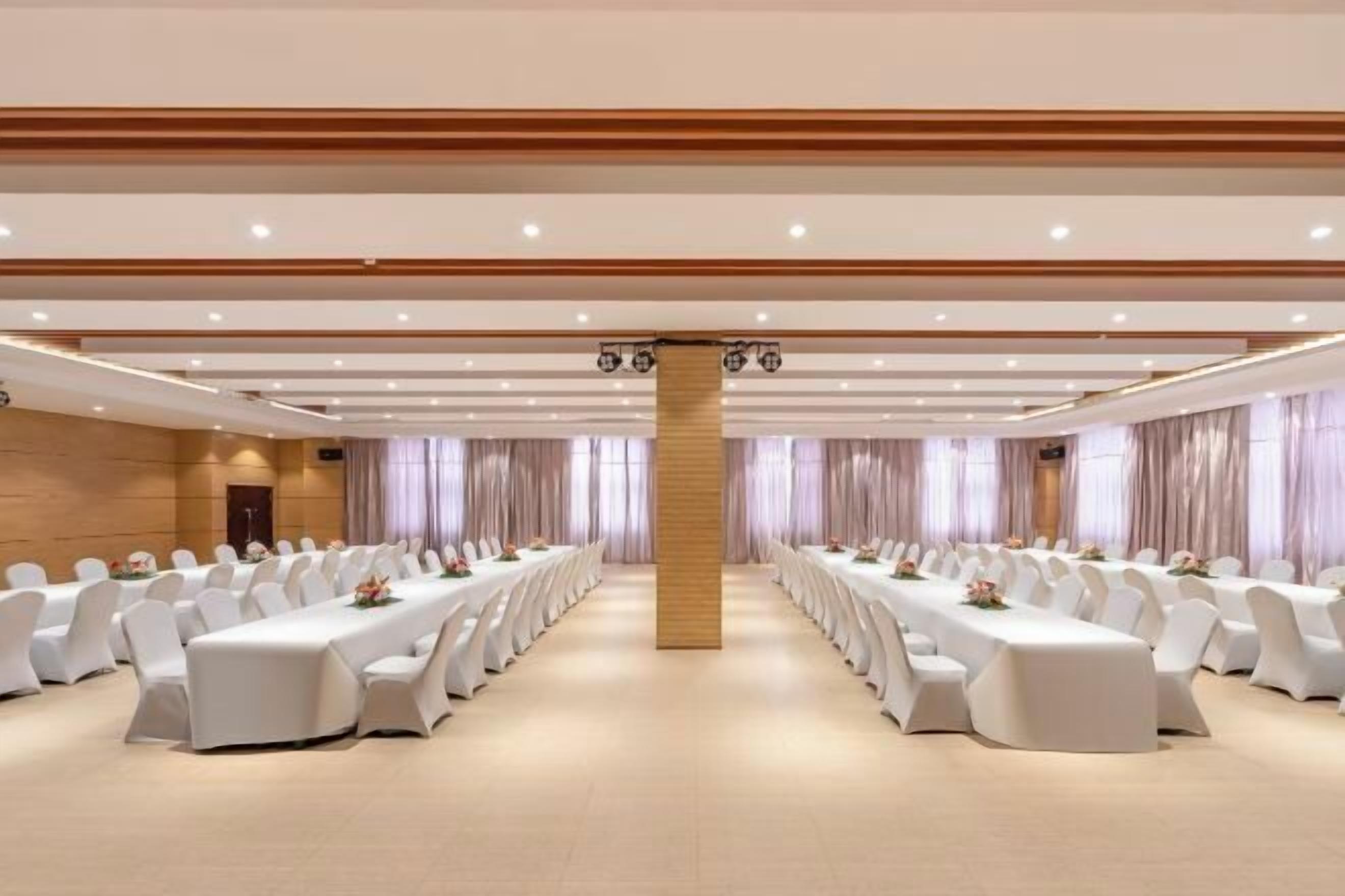 Un salón de conferencias con largas mesas y sillas blancas, un pilar de madera y un techo alto con iluminación empotrada.