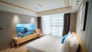 Una habitación de hotel moderna con una cama grande, televisión, sillón y ventana con cortinas.