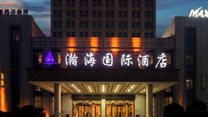 Entrada de hotel con señalización iluminada en un idioma extranjero, un área de acceso cubierto y un edificio alto al fondo.