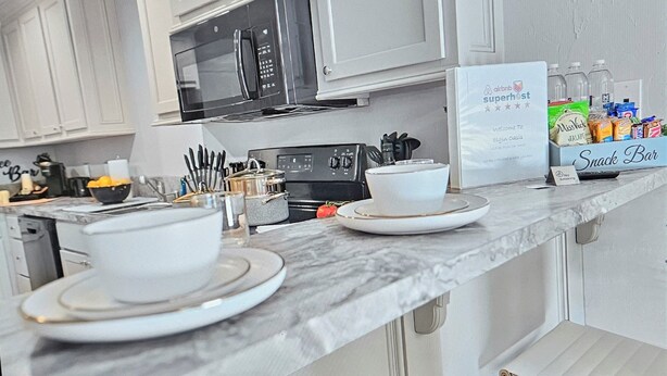 A kitchen with white cabinets, a microwave, a stove, and a snack bar with various food items. - Elgin Oasis Suite B (Elgin)