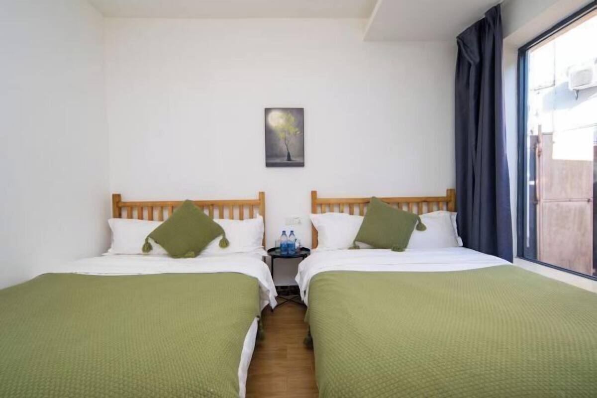 A hotel room with two beds, a small table with water bottles, a painting on the wall, and a window with curtains.
