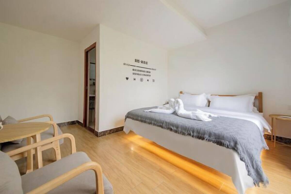 A hotel room with a bed, a small table, and a chair. There is a wall with a sign and a door.