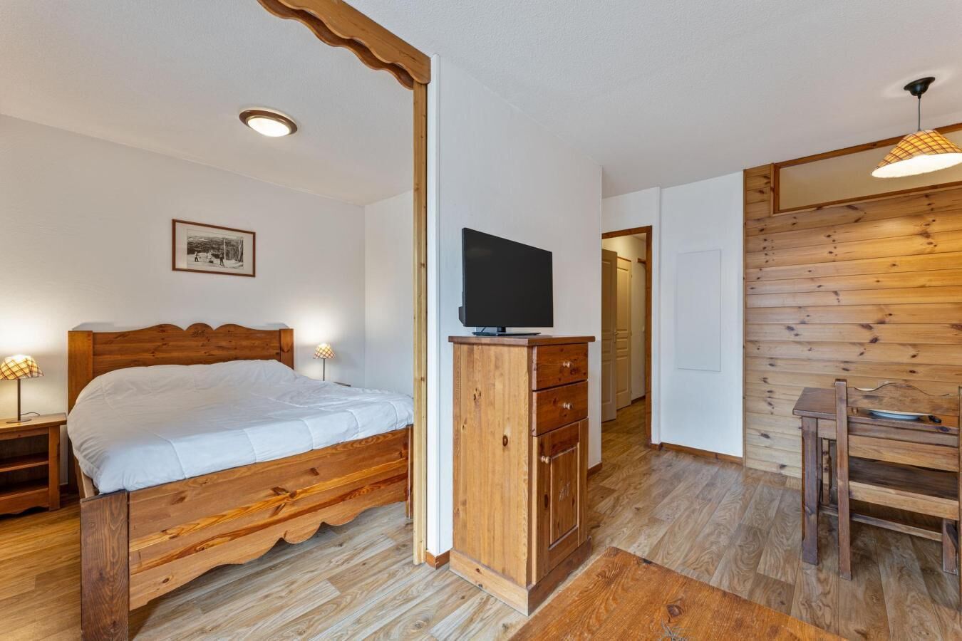 A wooden bed with a TV on a wooden cabinet, a wooden dining table, and a wooden wall with a picture frame.