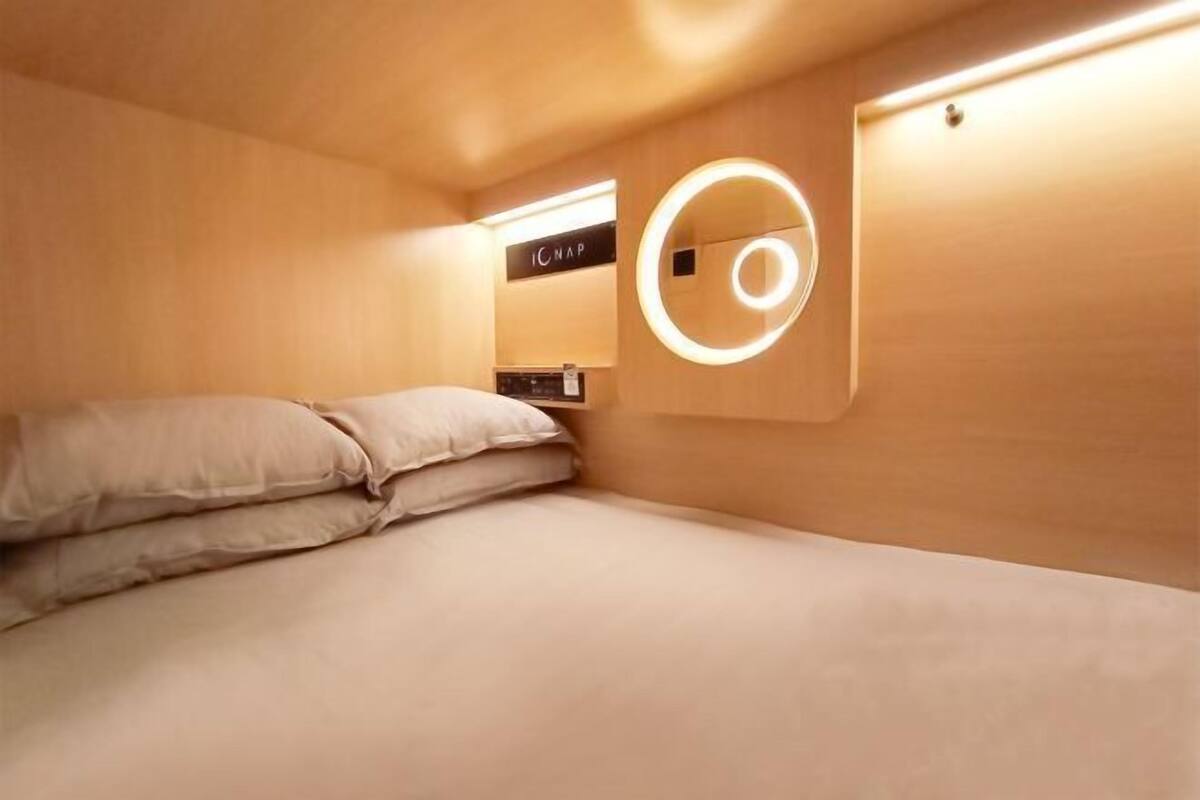 A compact, modern room with a bed, a circular light fixture, and a control panel on the wall.