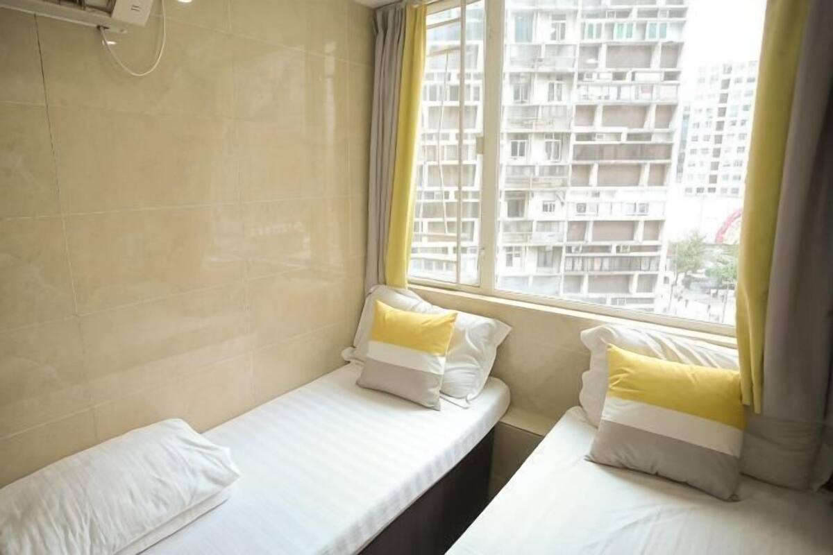 A hotel room with two single beds, a window with a view of a building, and a wall-mounted air conditioning unit.