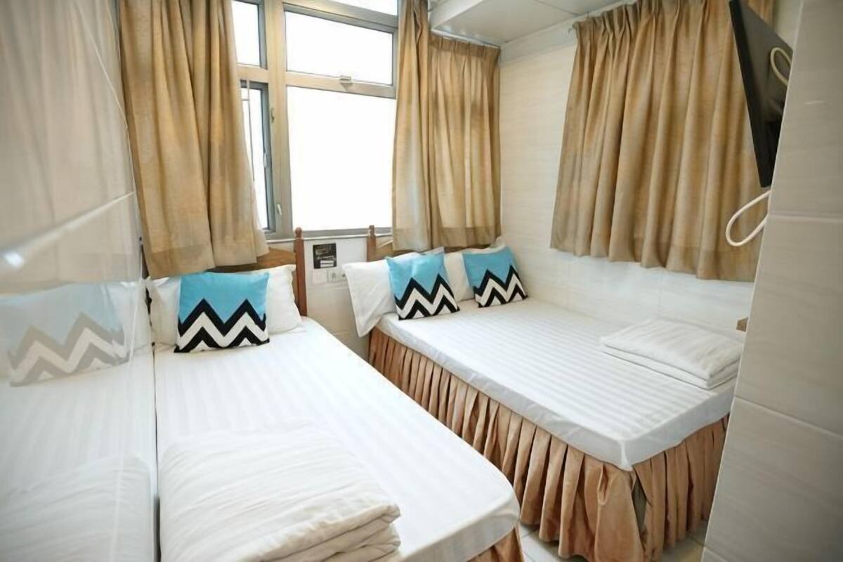 A room with two beds, beige curtains, a window, and a wall-mounted device.