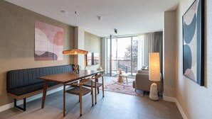 A modern hotel room with a wooden dining table, two chairs, a bench, a large window with curtains, a sofa, a lamp, and abstract wall art. - 3-bedroom Holiday Home in Den Haag (The Hague)