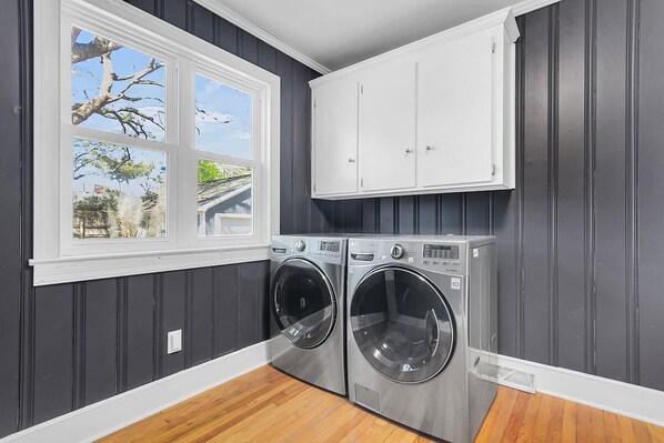 A laundry room with two front-loading washing machines, a window with a view of trees and a house, and white cabinets above the machines.