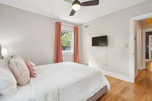 A bedroom with a bed, a ceiling fan, a TV mounted on the wall, and a window with curtains.