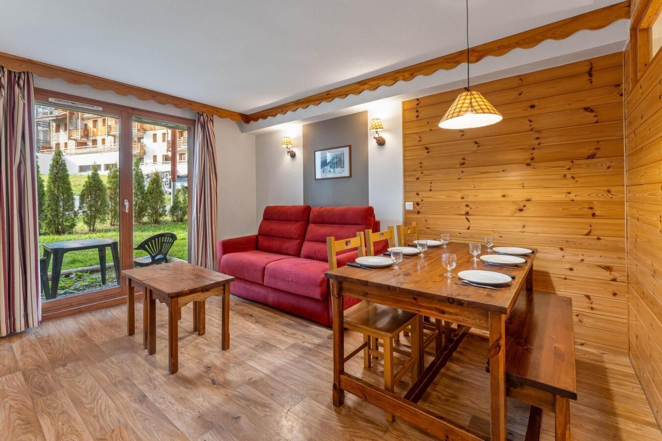 A cozy room with a wooden dining table set for four, a red sofa, and a view of a green garden through large windows.