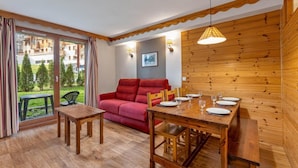 A cozy room with a wooden dining table set for four, a red sofa, and a view of a green garden through large windows. - 40m from ski slopes, fitness, swimming pool, sauna, tv, ski locker, 41m², Les Orres (Les Orres)