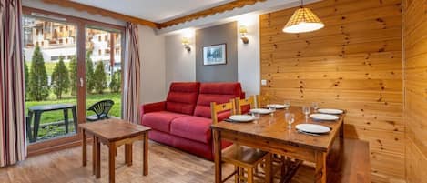 A cozy room with a wooden dining table set for four, a red sofa, and a view of a green garden through large windows.