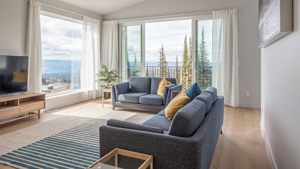 A modern living room with a large window, grey sofas, a flat-screen TV, and a wooden coffee table. - Brand new, full duplex sleeps 22 - hot tub, laundry, sauna, BBQ w/ views (Silver Star Mountain)