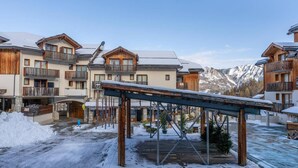 Un espace extérieur enneigé, avec des bâtiments en bois, une allée couverte et une chaîne de montagnes en arrière-plan.