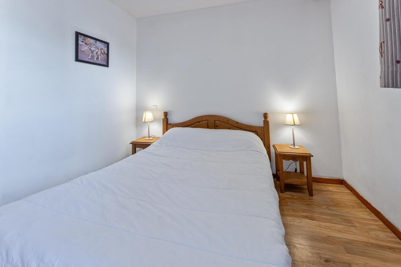Un lit simple avec une literie blanche, dans une pièce au plancher en bois et aux murs blancs. Deux tables de chevet avec des lampes sont placées de chaque côté du lit.