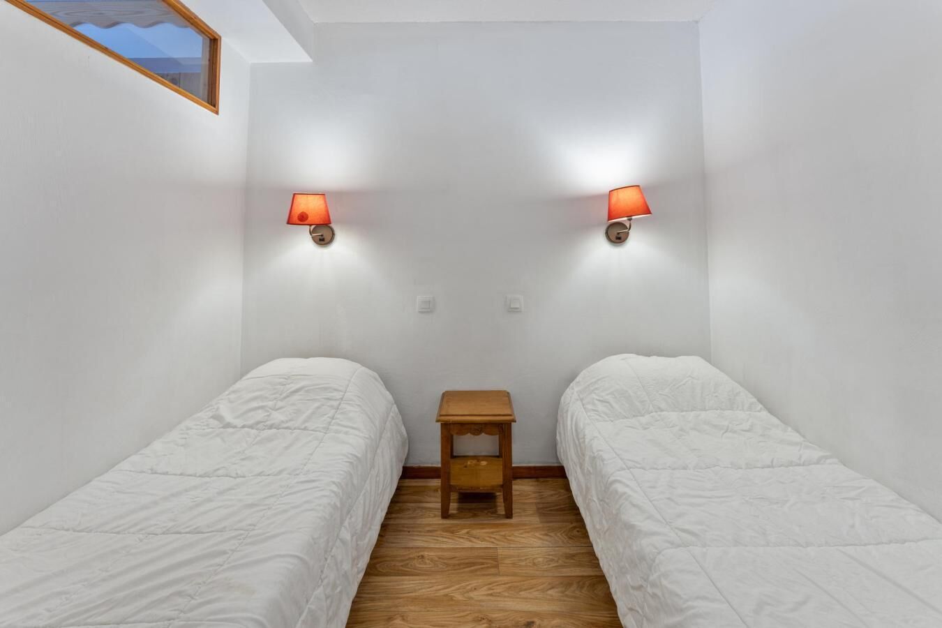 A small, single-room bedroom with two beds, a wooden nightstand, and two wall-mounted lamps.