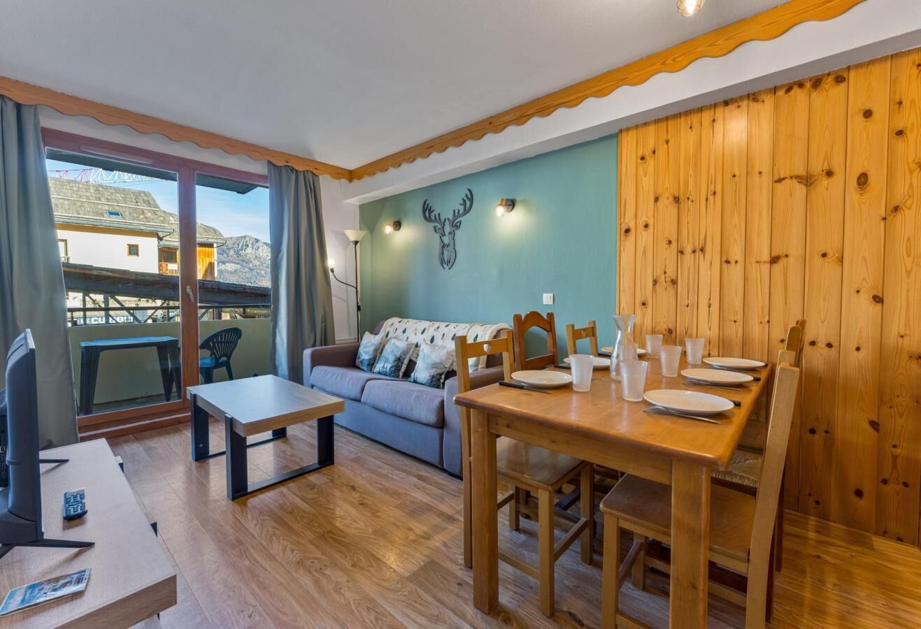 A cozy living area with a sofa, a wooden dining table set for four, and a balcony with a view of the mountains.