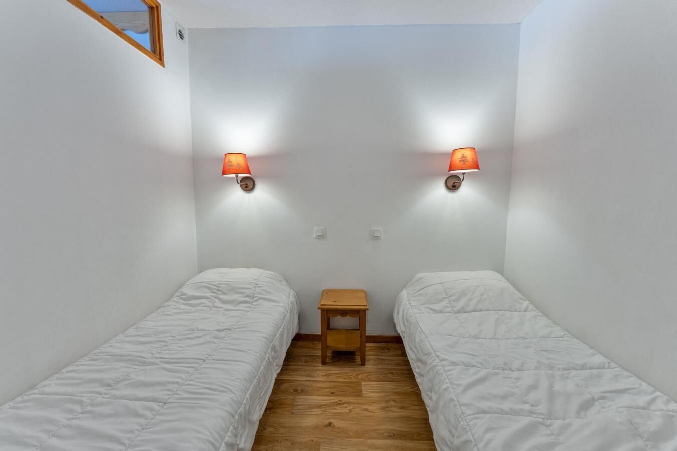 A small, single-room accommodation with two single beds, a small wooden table, and two wall-mounted lamps.