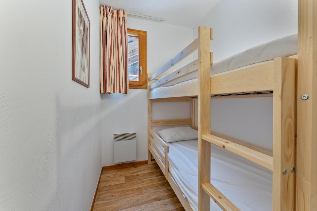 A narrow room with wooden bunk beds, a radiator, and a window with curtains.