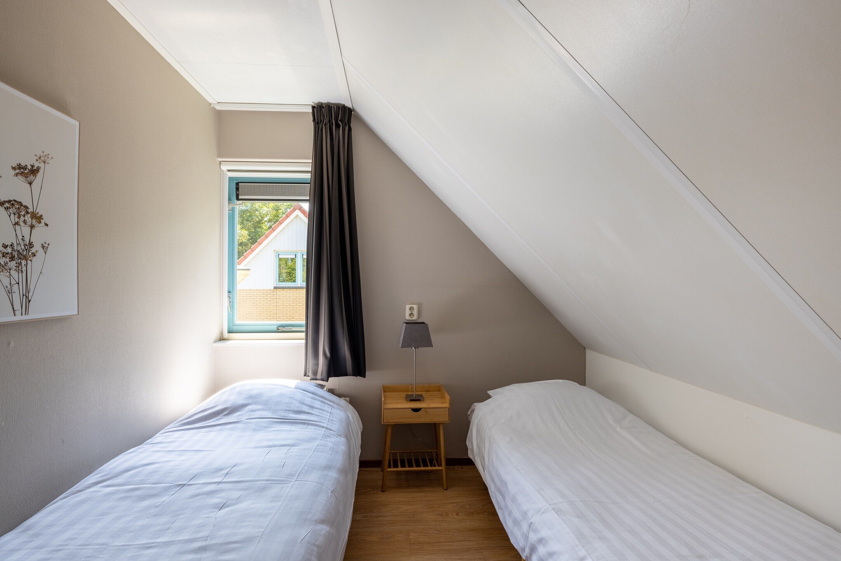 A small, well-lit room with two single beds, a nightstand with a lamp, a window with curtains, and a view of a building outside.