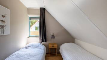 A small, well-lit room with two single beds, a nightstand with a lamp, a window with curtains, and a view of a building outside.