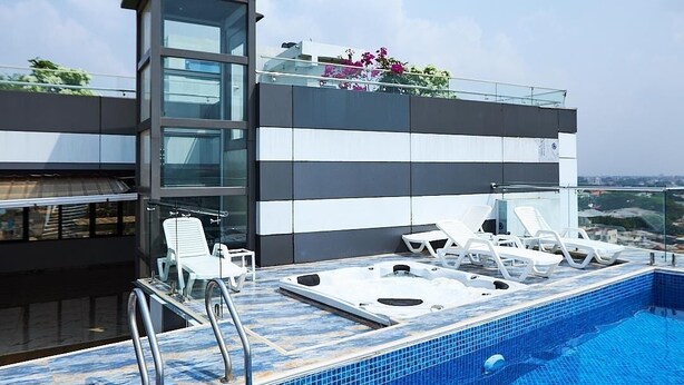 A rooftop pool area with lounge chairs and a hot tub, surrounded by a modern building with large windows and a view of the city. - Luxe Apartment with Rooftop Pool & Gym near the Airport. (Accra)