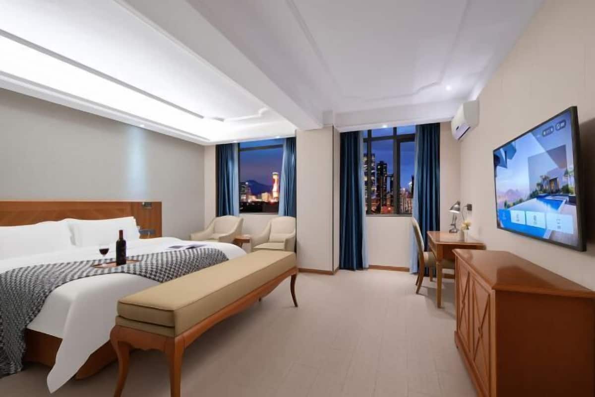 Hotellihuone, jossa on sänky, penkki, työpöytä, televisio ja näkymä kaupungin yömaisemaan.