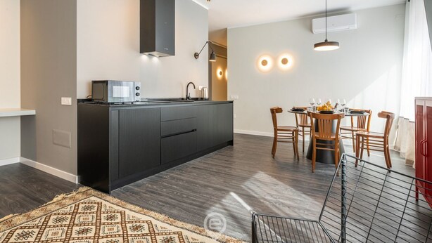 A modern kitchen with dark cabinetry, a built-in microwave, and a dining area with wooden chairs and a table. - Saint Margaret Apartments (Cagliari)