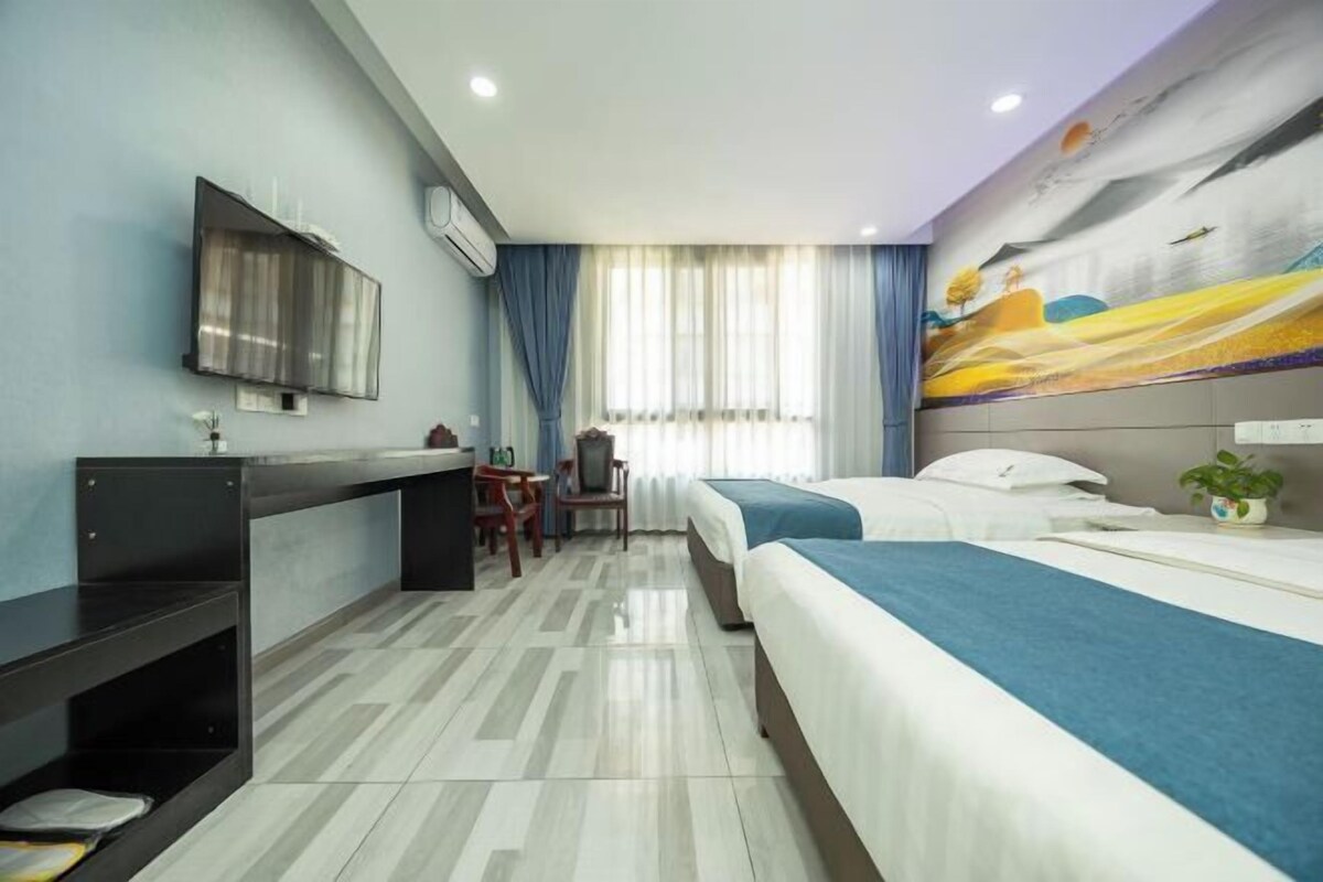 Habitación de hotel con dos camas, un escritorio, una silla, un televisor y un ventanal grande con cortinas.