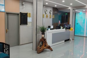 A reception area with a reception desk, a wooden plant stand, a wall clock, and a digital screen. - Yinlilai Business Hotel (Tianquan Hospital of Traditional Chinese Medicine Branch) (Tianquan)