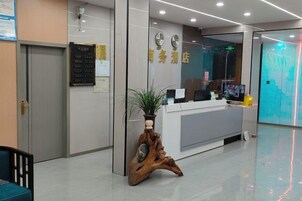 Un área de recepción con mostrador, un soporte de madera para planta, un reloj de pared y una pantalla digital.