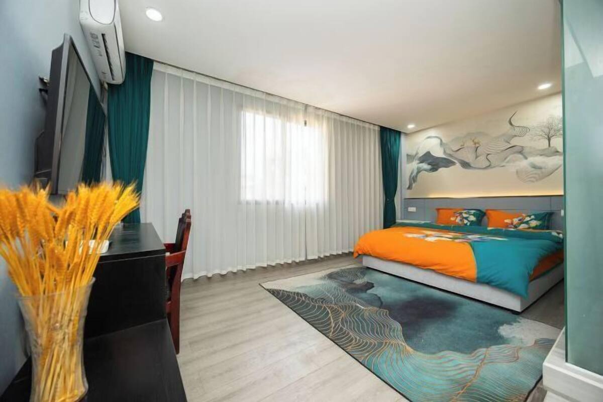 A modern hotel room with a bed, a desk, and a chair. There is a window with curtains, a wall-mounted air conditioner, and a decorative wall piece.
