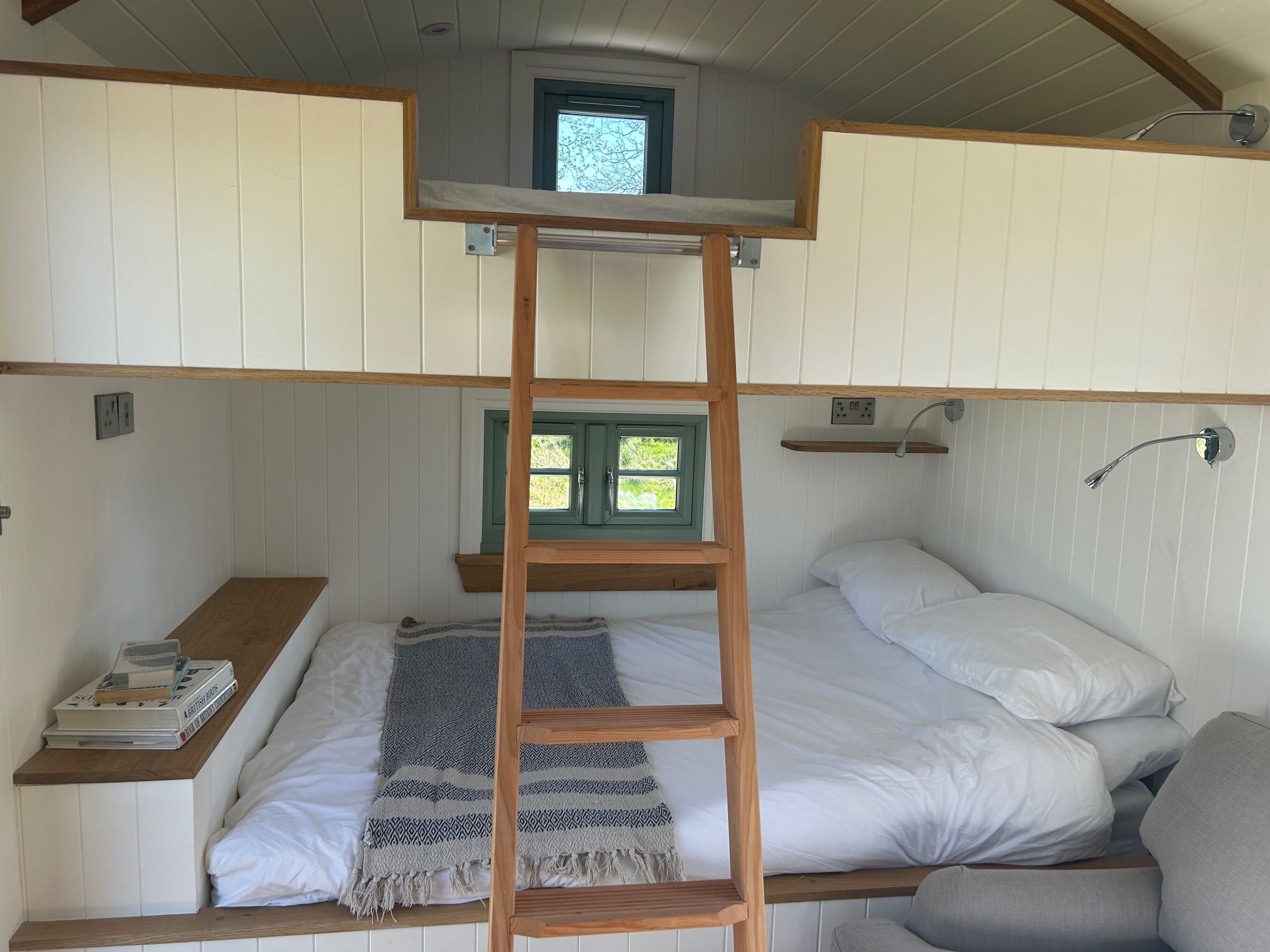 Uma cama beliche com escada de madeira, uma pequena janela e uma mesa de cabeceira com livros.